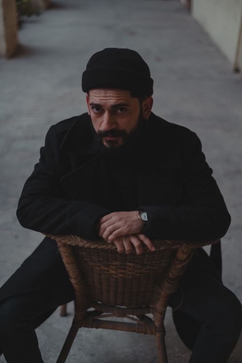 Free Bearded man in black outfit and hat looking at camera while sitting backwards on wooden chair against blurred background Stock Photo