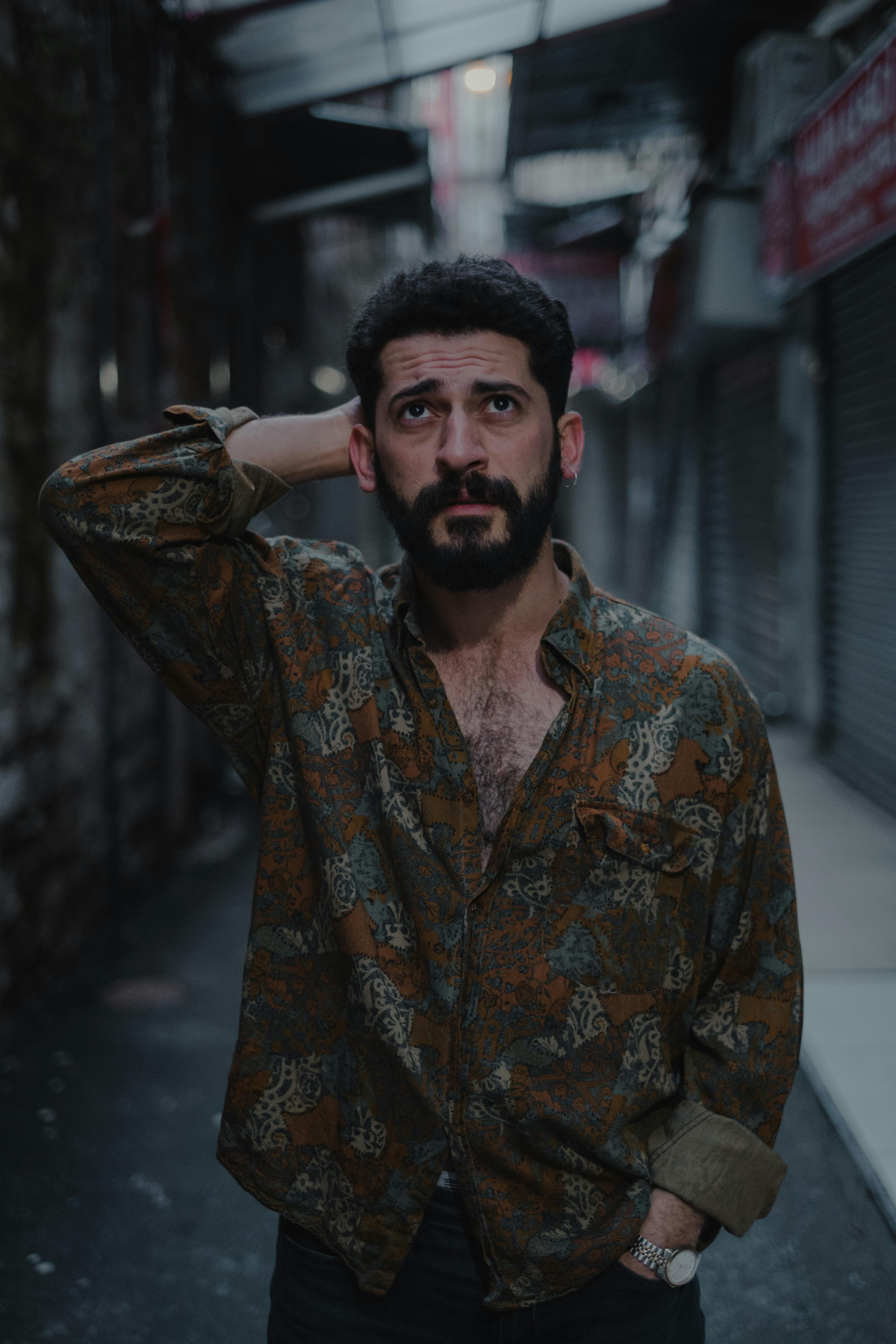 Ethnic bearded man in casual outfit looking up with hand on head while standing on asphalt road on backstreet