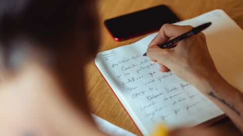A person writes in a notebook at a wooden desk with a phone nearby.