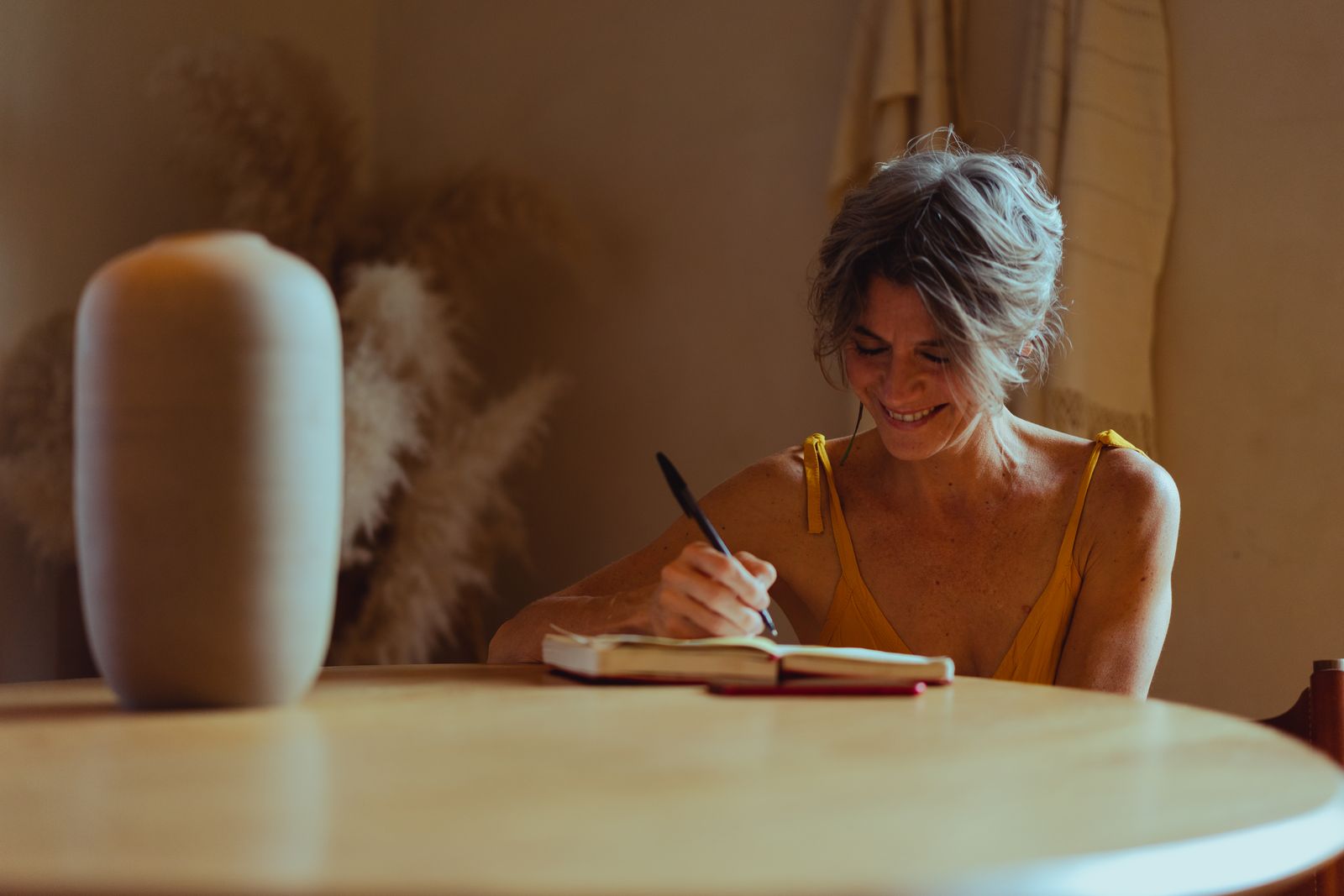 Smiling woman with short gray hair writing in a notebook at a round wooden table in warm light
