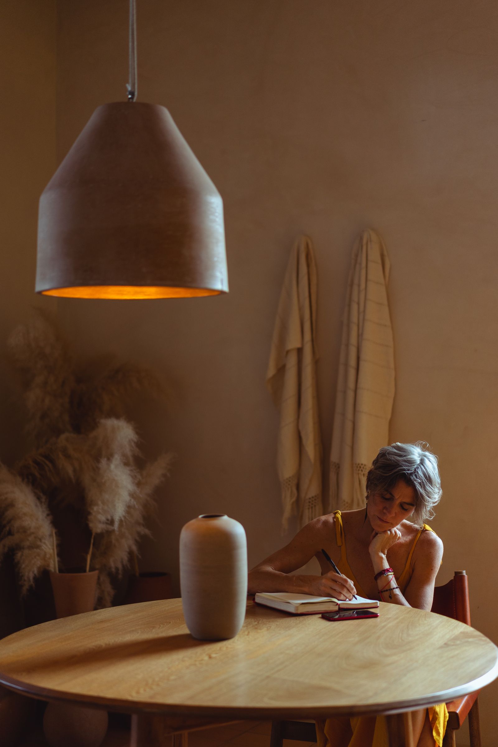 An older woman writing at a round wooden table beneath a warm hanging lamp