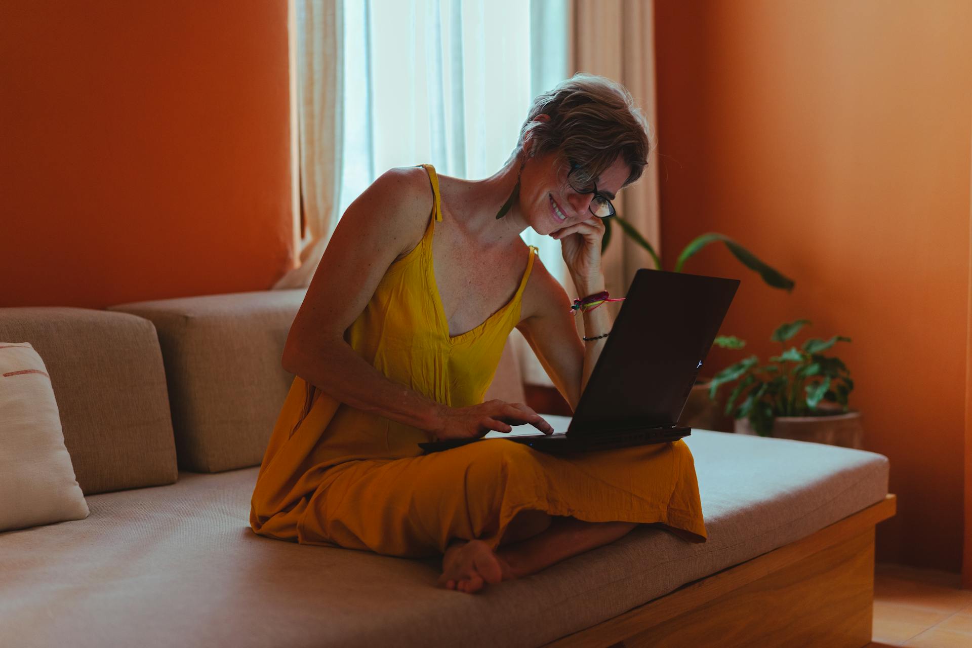 Senior woman sitting on a sofa, happily working on a laptop from home, showcasing positive aging and modern tech.