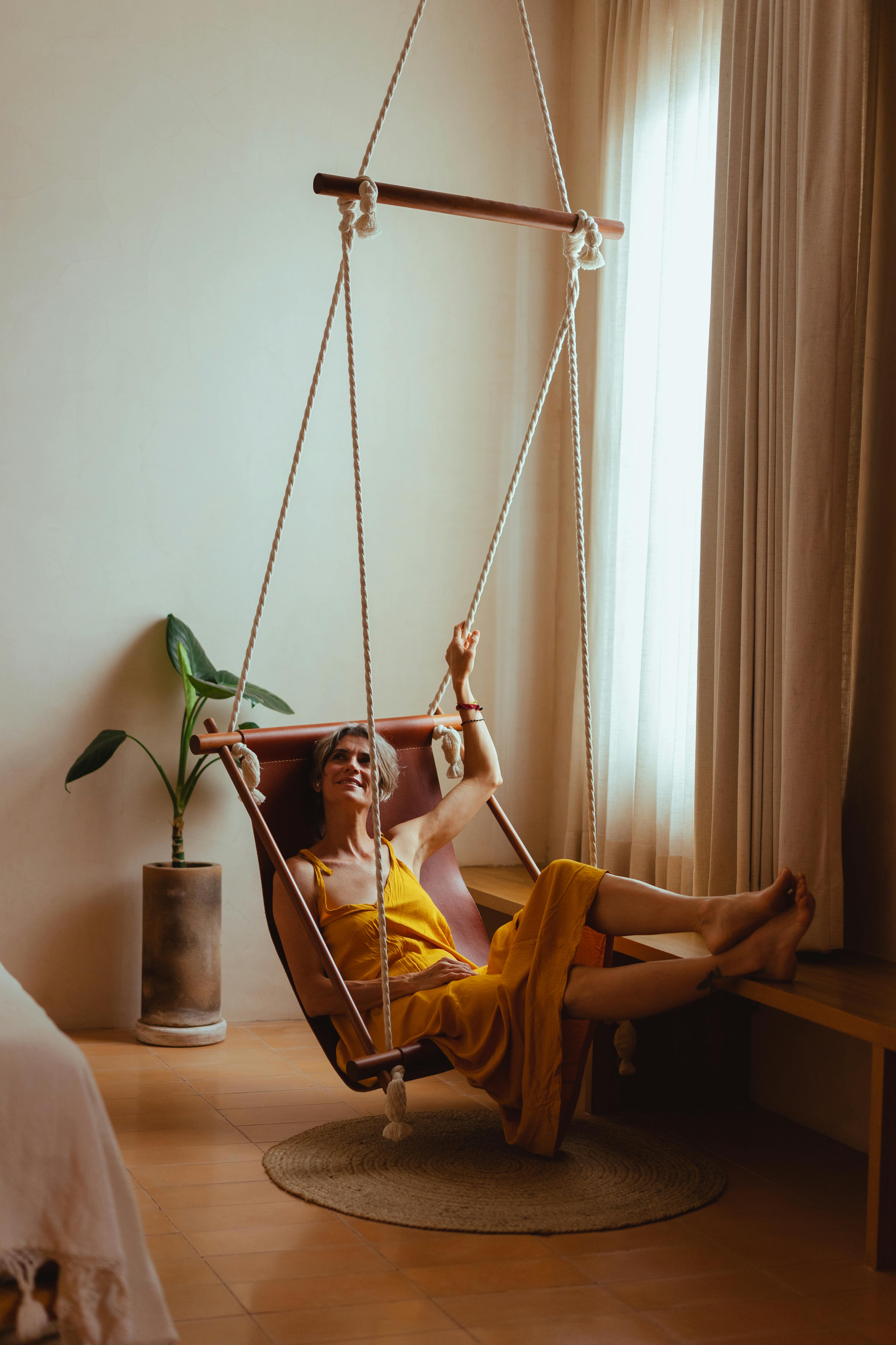 An elderly woman in a yellow dress smiles while relaxing in a hammock chair indoors.