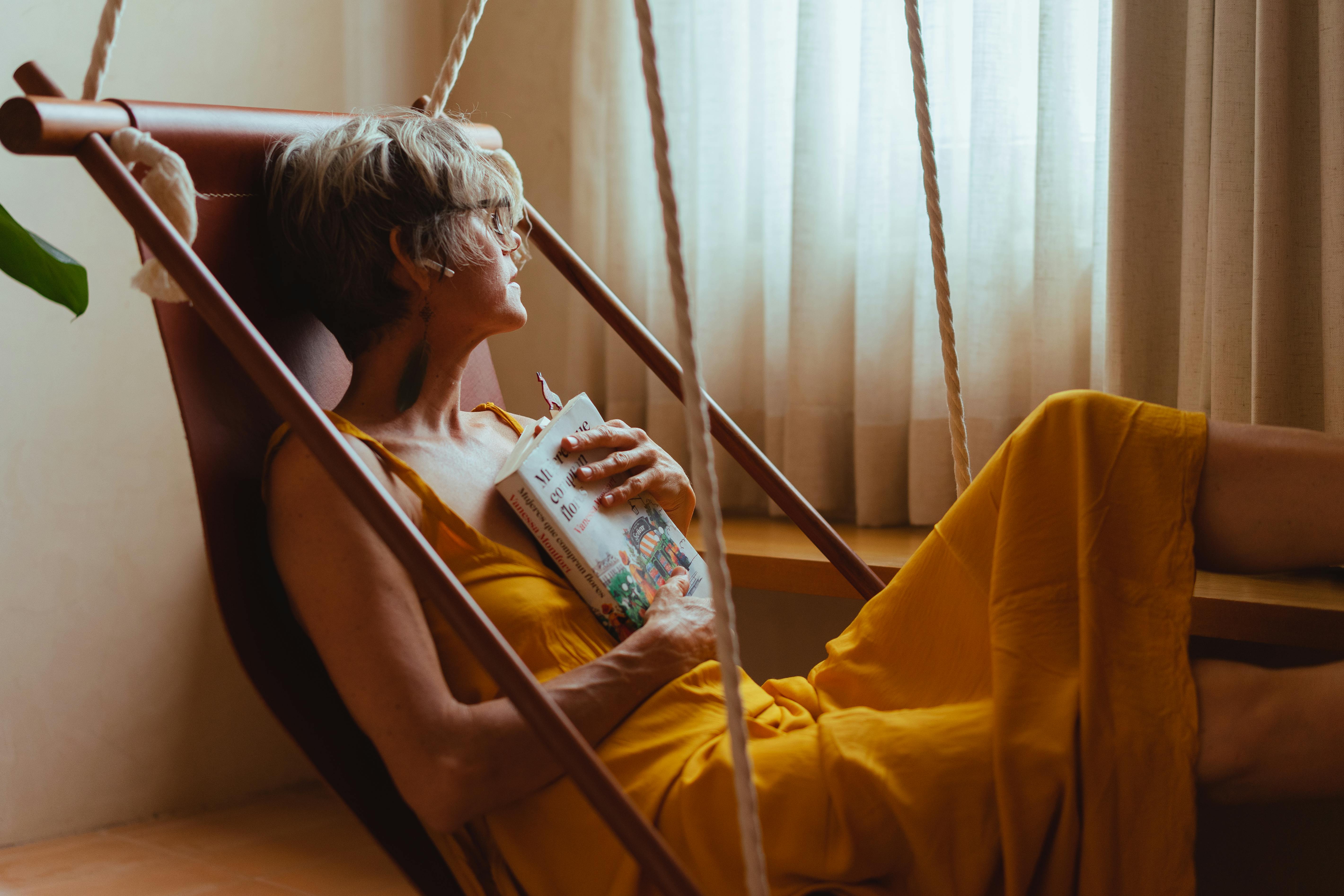 Senior woman in a hammock, embracing a book, enjoying leisure time indoors.
