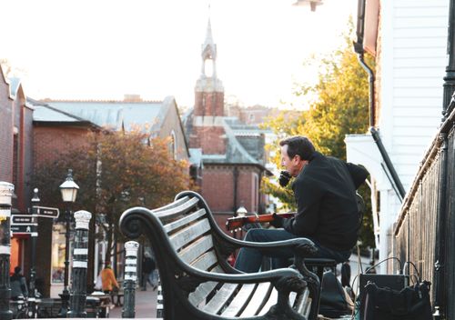 Free Man sitting on a bench playing guitar outdoors in a quaint urban street setting. Stock Photo