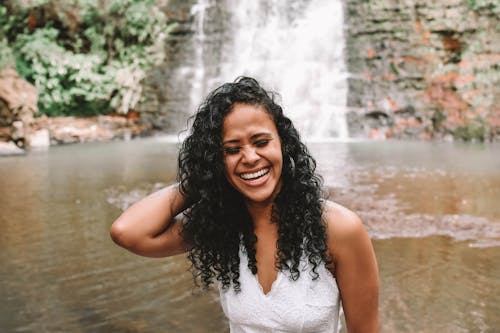Free Laughing African American female with closed eyes standing near river against streaming waterfall falling from rocky cliff in wild nature Stock Photo
