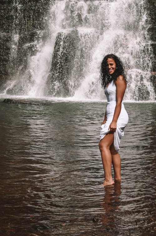 Free Full body of cheerful African American female in white outfit standing in river near rapid waterfall flowing through rocky cliff Stock Photo