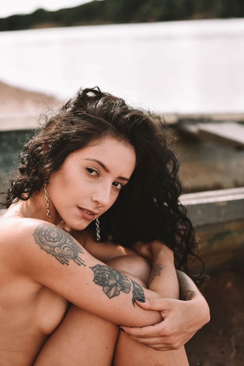 Free Charming nude female with tattoos looking at camera while sitting on shore with wooden boat near river on blurred background Stock Photo