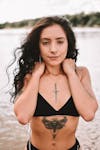 Free Attractive female with curly hair and tattoos wearing black swimwear and looking at camera while standing near water on blurred background Stock Photo