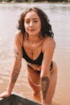 Free Glad slim female with tattoos wearing black swimsuit and looking at camera while standing in river while leaning on boat Stock Photo