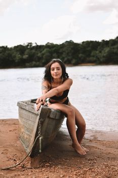 Full body of barefoot tattooed female in swimwear looking at camera while sitting on shabby wooden vessel on beach near river