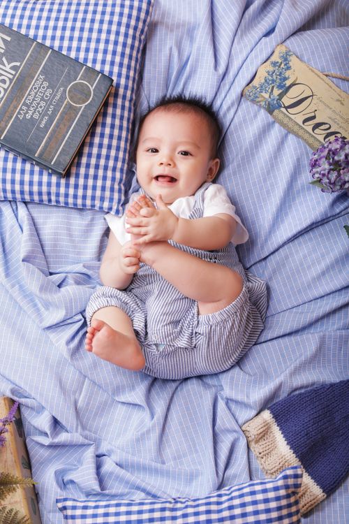 Free Cute baby boy smiling and holding his foot while lying on blue checkered bed linens, surrounded by books and fabric. Stock Photo