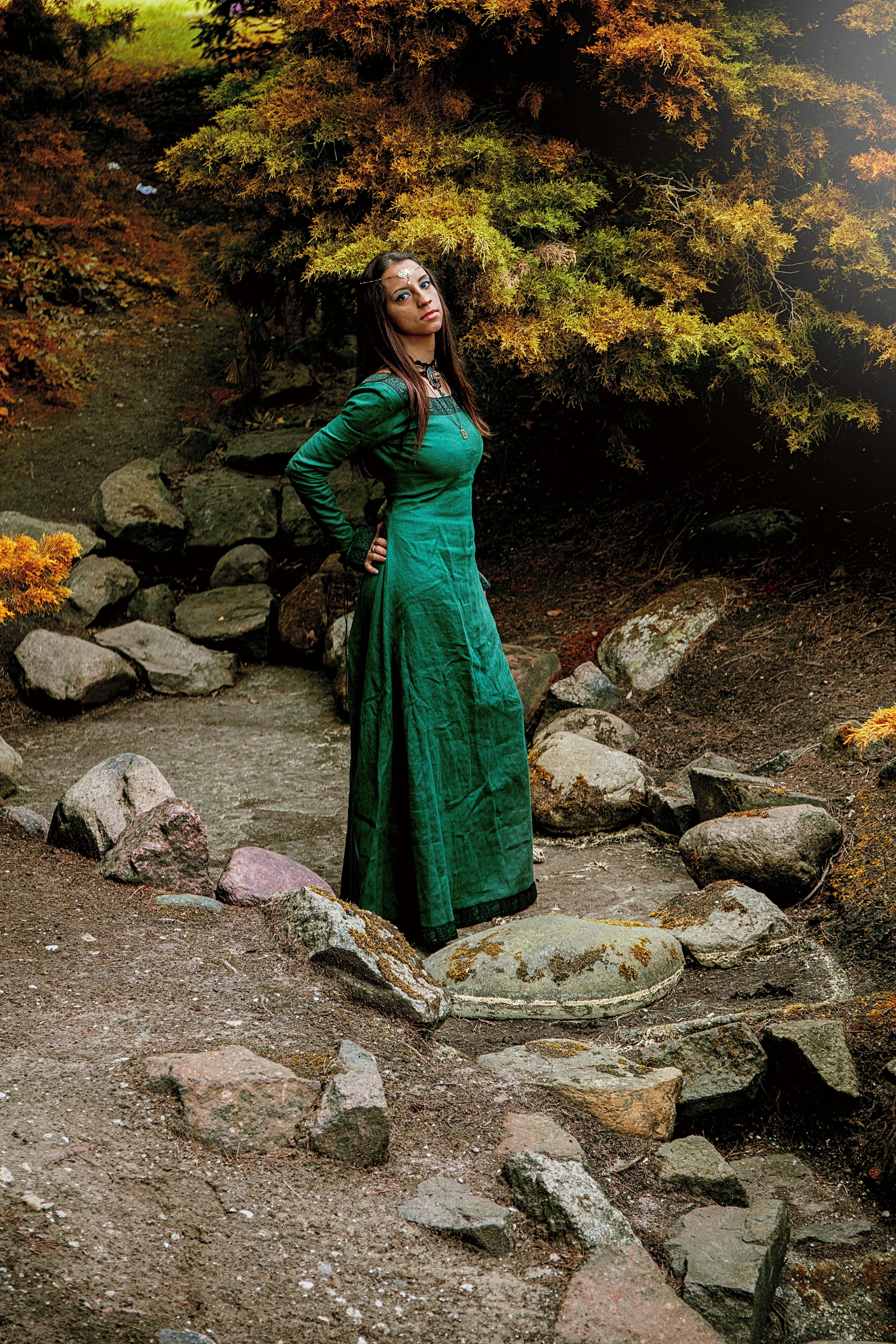 A woman in a green dress poses gracefully in a forest setting, portraying a fantasy theme.