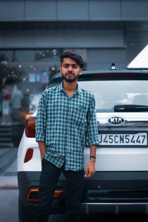 Free Confident young man in plaid shirt standing by car in urban area, embodying modern lifestyle. Stock Photo
