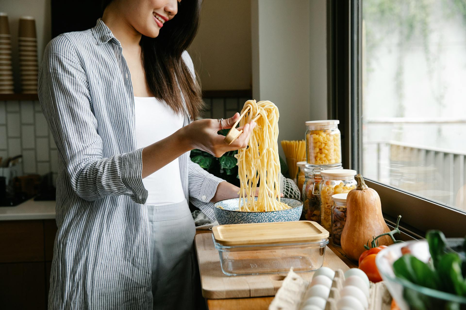 Side view of cheerful female with long cooked spaghetti on tongs standing in kitchen while preparing delicious pasta at home