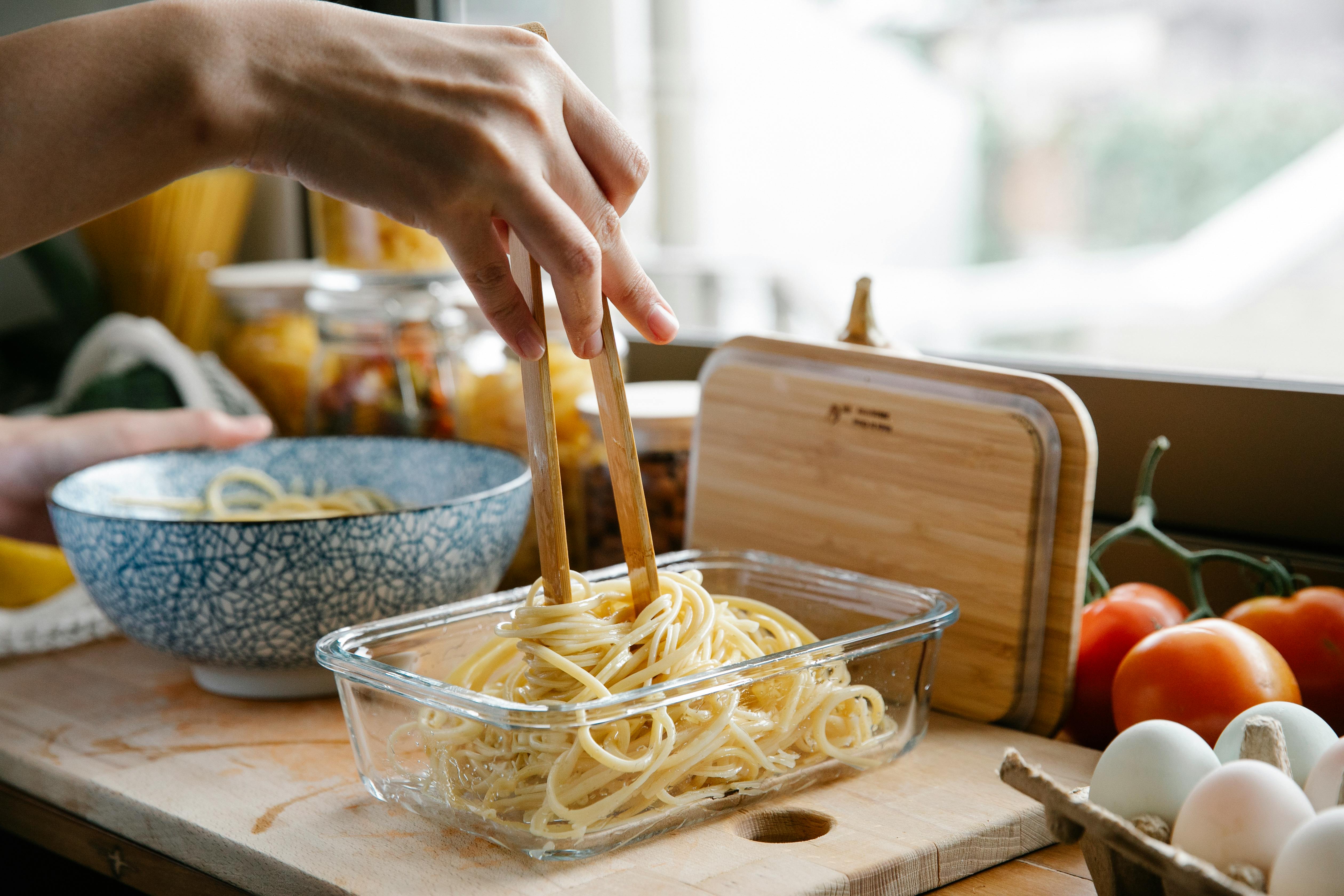 Unrecognizable female cook with tongs standing at counter with glass container of fresh pasta while cooking in kitchen on blurred background