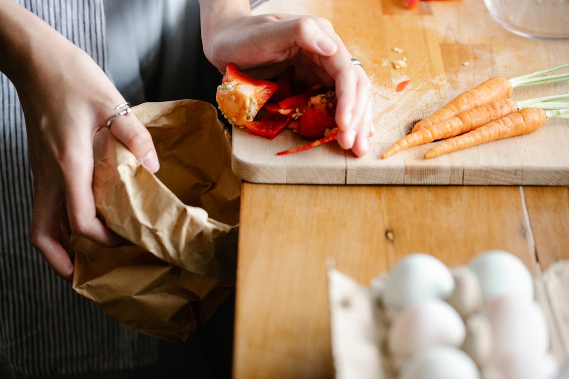 Free Unrecognizable female putting seeds of red bell pepper from cutting board into carton package while cooking at counter with carrots and eggs Stock Photo