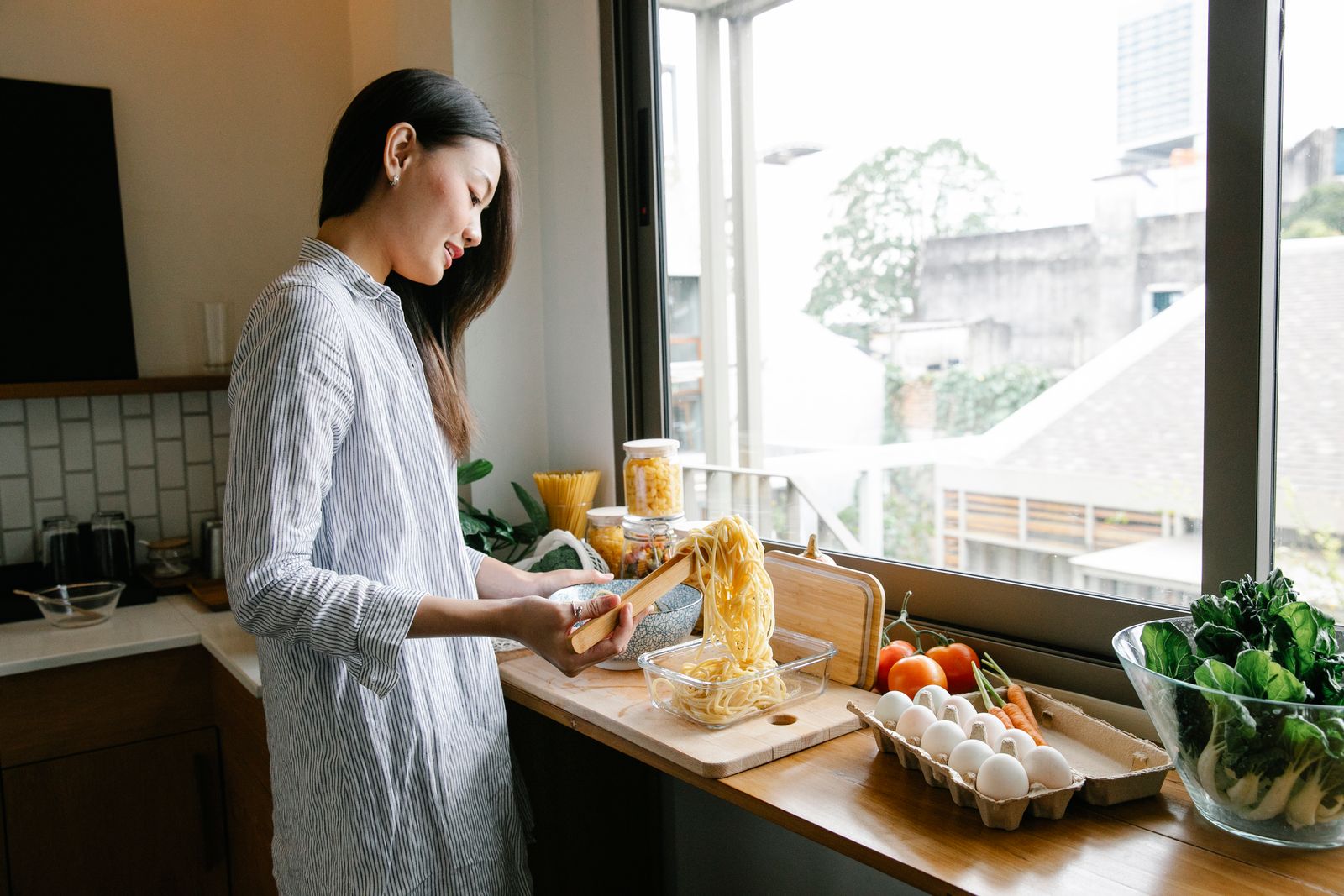 Side view of Asian female cook putting long spaghetti into glass container while standing at counter with various ingredients in kitchen