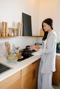 Side view of concentrated Asian female cook standing near cooker with saucepan while cooking tasty pasta in kitchen at home