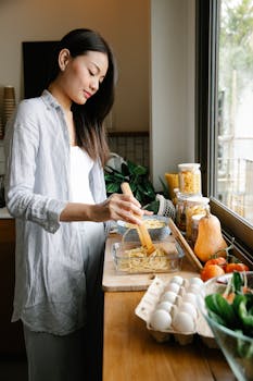 Side view of concentrated Asian female cook putting cooked pasta into glass container while cooking lunch in kitchen at home