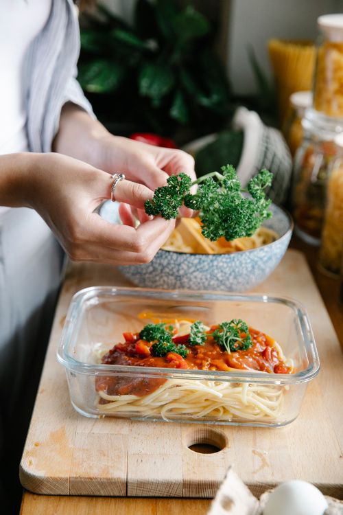 Free Unrecognizable female cook adding herbs into container with spaghetti and bolognese sauce while standing at counter and cooking in kitchen Stock Photo