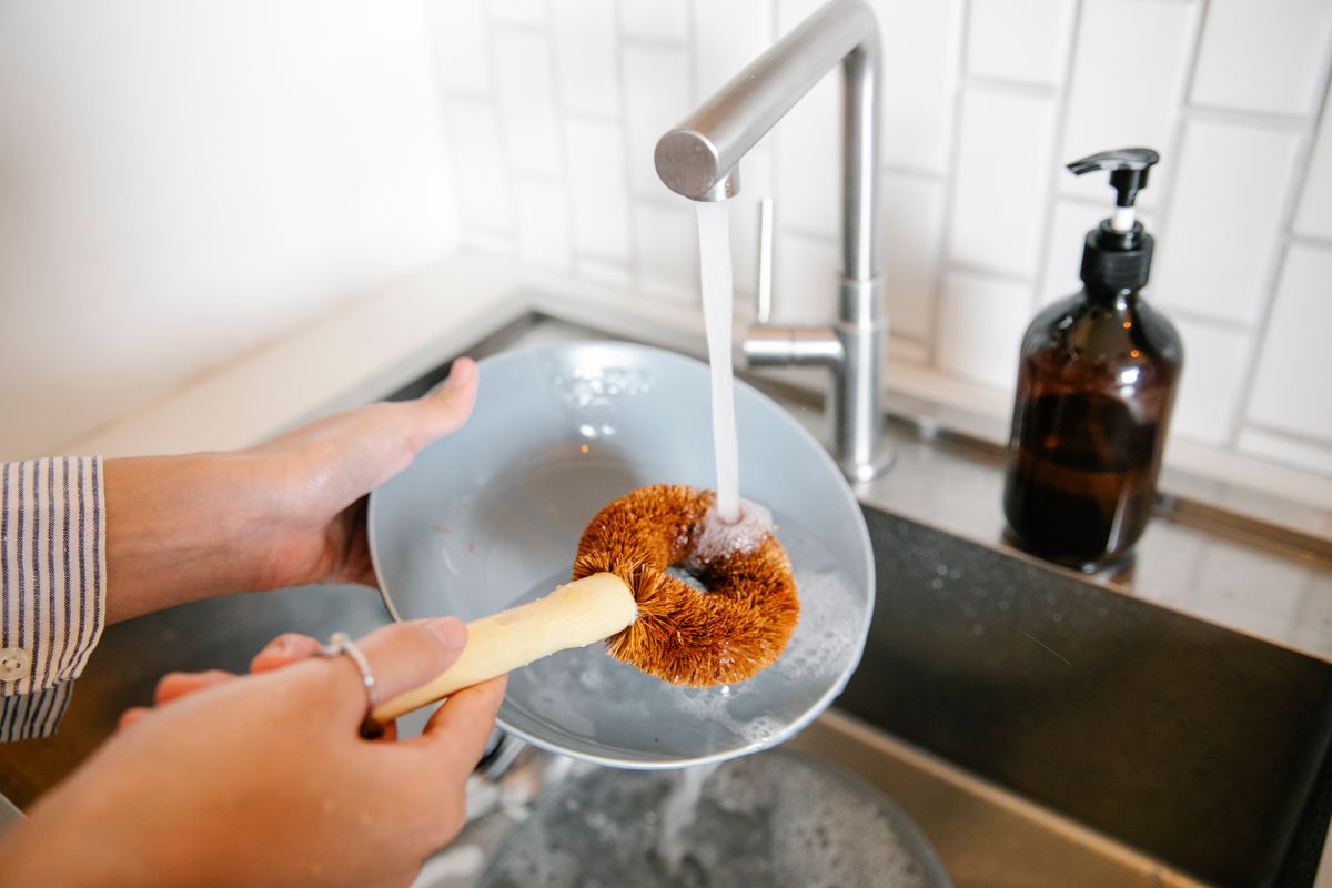 Close-up of hands washing a dish with a brush under running water at a kitchen sink