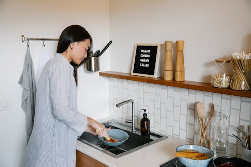 Side view of Asian female with brush washing dishes at sink while doing housework in light kitchen with supplies at home