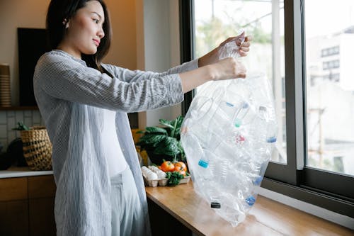Side view of positive Asian female with transparent bag full of plastic bottles for recycling standing in light kitchen at home