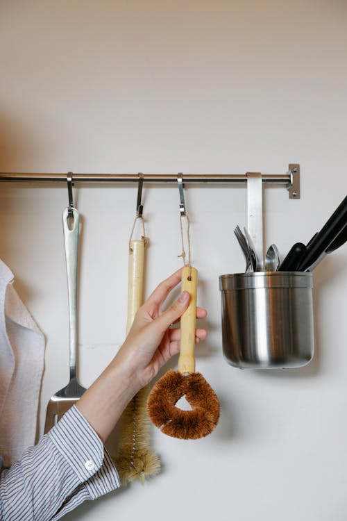 Unrecognizable female hanging brush for washing dishes on metal rack on wall with various equipment in light kitchen at home
