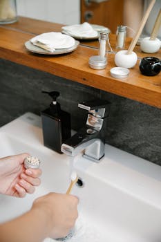 Hand applying natural toothpaste on a bamboo toothbrush near bathroom sink.