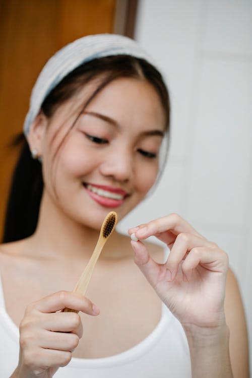 Charming Asian female looking at wooden toothbrush and toothpaste tablet in hands while standing in light bathroom during morning routine