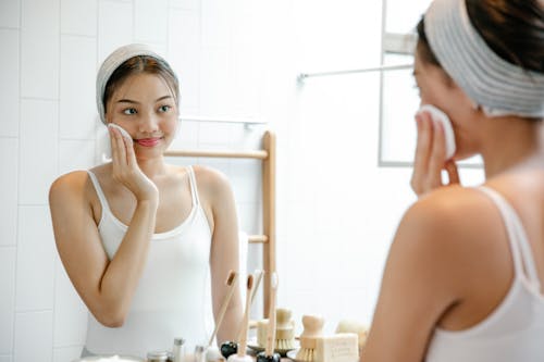 Smiling young Asian female looking at reflection in mirror while cleaning face skin with cotton pad during daily morning routine in bathroom