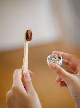 Anonymous female with wooden toothbrush and small jar of toothpaste tablets standing in light room on blurred background during hygienic routine
