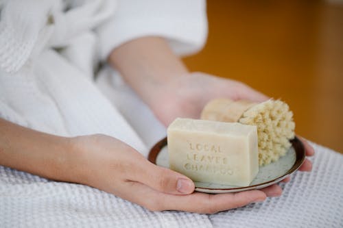 Anonymous female with soap and brush for skincare treatment on plate sitting in light room during daily procedure at home