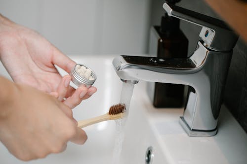 Crop anonymous person with toothpaste tablets and ecological toothbrush under aqua flowing from washbasin tap in bathroom