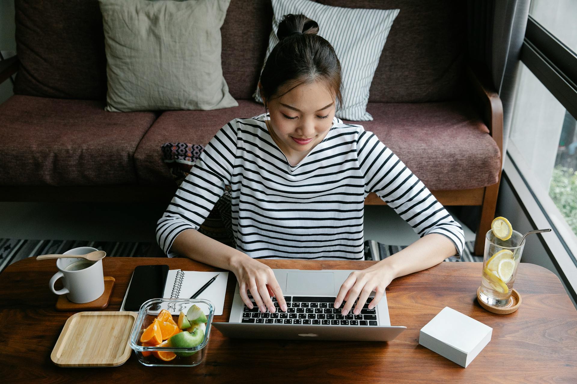 Young Asian woman using a laptop at home with fresh lemonade and fruit, working in a calm environment.