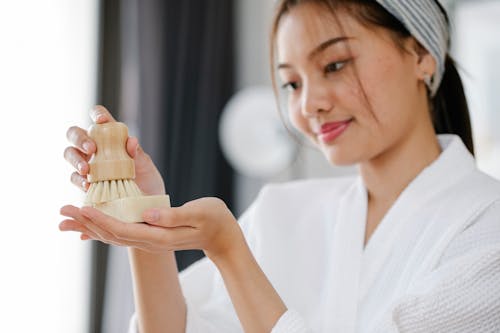 Charming Asian female in white bathrobe with soap and body brush for skincare treatment standing in room on blurred background