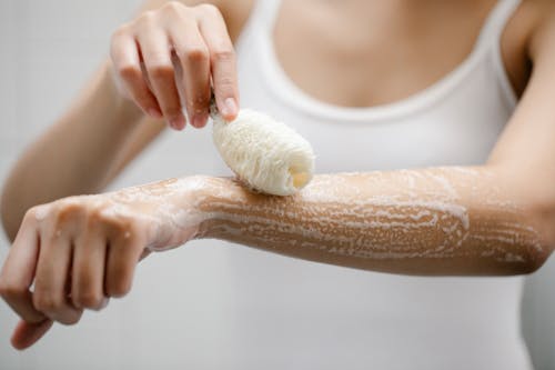 Unrecognizable female with shower sponge washing arm with foam while standing in light bathroom during daily skincare routine at home
