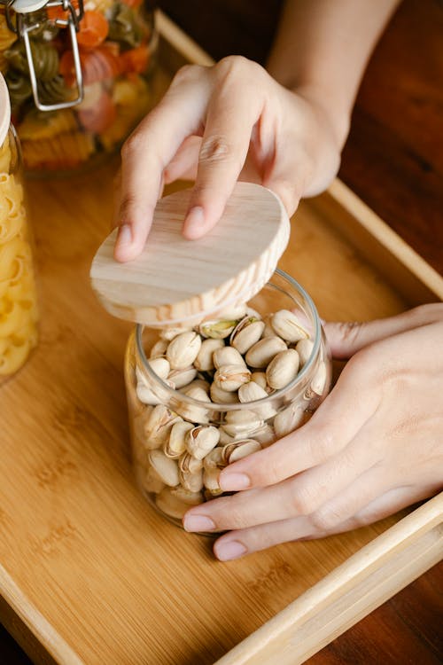 From above anonymous person opening glass jar of pistachios with wooden lid on table