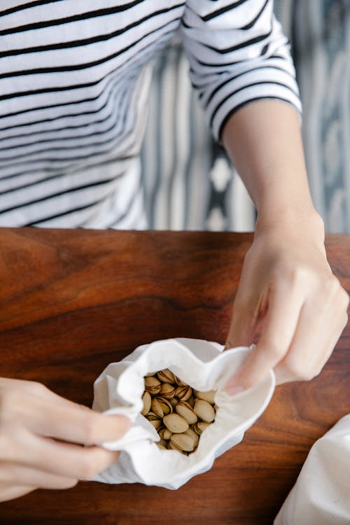 From above crop unrecognizable person in casual clothes opening small white recycling bag with pistachios while standing at wooden table