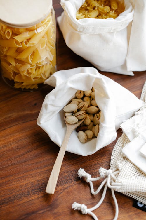 From above pistachios in white recycling bag with wooden spoon and pasta near penne in glass jar and rotini in white recycling bag on wooden table