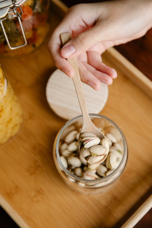 High angle of crop person holding wooden spoon with pistachios under glass jar on wooden table