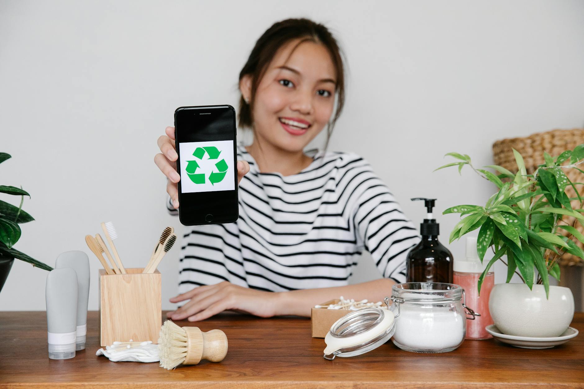 Smiling Asian woman demonstrating recycling symbol on smartphone while sitting at table with eco friendly cosmetic products and looking at camera