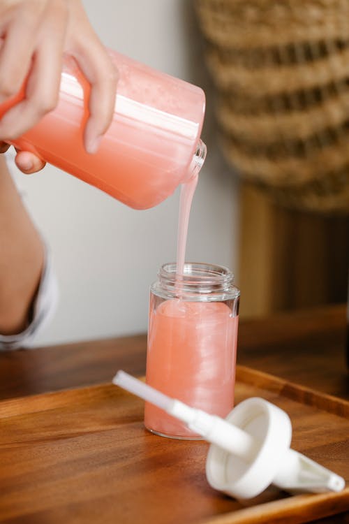 Crop anonymous person standing at table with opened soap dispenser and pouring peach coloured soap into bottle