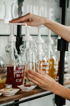 Crop unrecognizable person pouring red soap into plastic bottle while pressing large dispenser near white saucers and glass jars on shelf