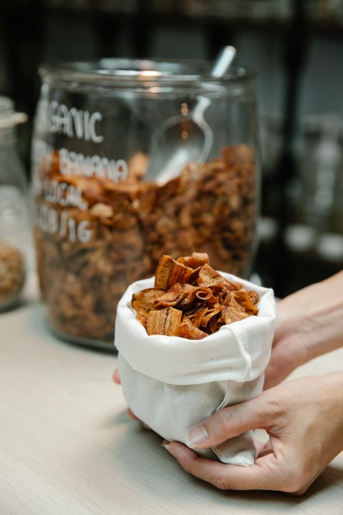 Free Close-up of hands holding a sack of organic banana chips with a glass jar in the background. Stock Photo