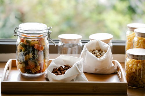 Free Glass jar with rotini pasta placed on wooden tray near ECO friendly bags with almonds and pistachios in light room near window on blurred background Stock Photo