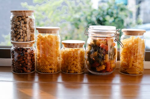 Glass jars filled with assorted types of uncooked pasta and pistachios with almonds placed on wooden table near window in light room