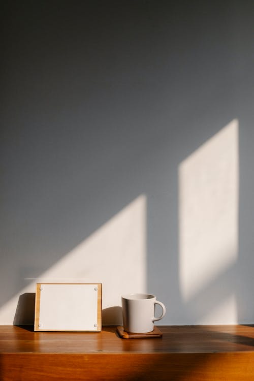 Empty white paper on notice board placed on wooden table near cup at wall in light room with bright sunlight