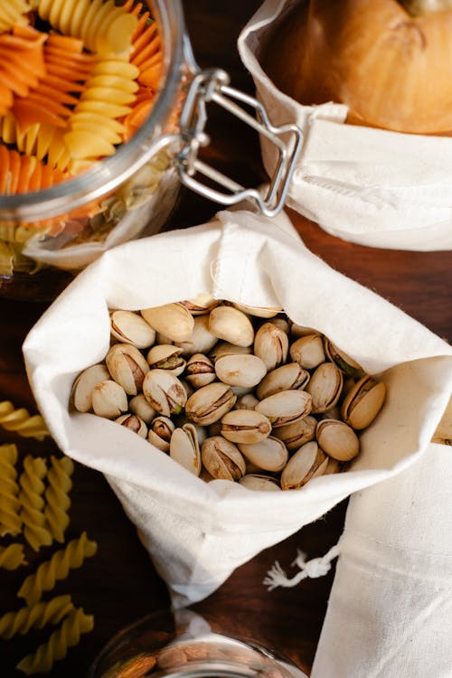 Top view of ECO friendly sack full of pistachios placed on table near raw pasta and ripe pumpkin in light room